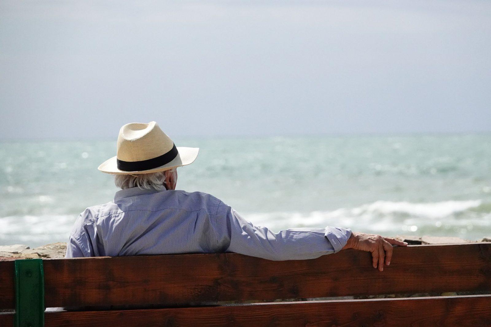 starszy pan w kapeluszu, siedzi na ławce i patrzy na morze.